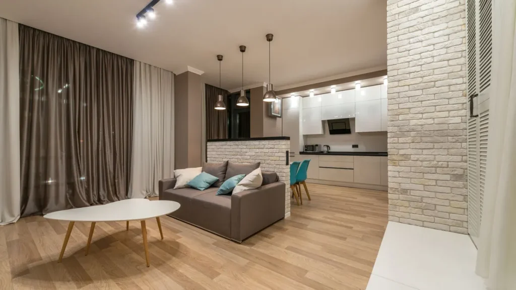 Woonkamer met lichte houten vloer, grijze bank en open keuken op de achtergrond